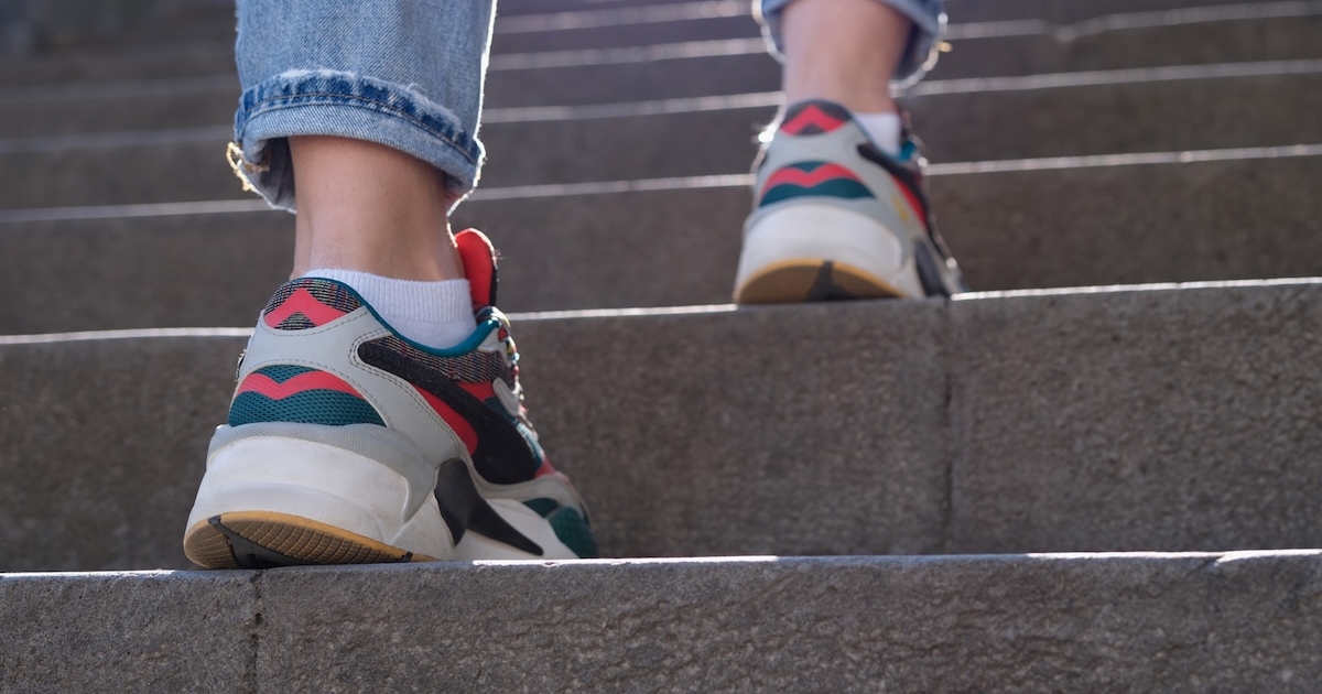 Close-up from behind of feet in colorful sneakers and cuffed jeans walking up grey stone stairs in bright sunlight